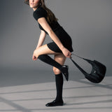Woman in black outfit with knee-high socks and a black handbag on a gray background thumbnail