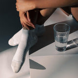 A person sitting on white geometric steps wearing white crew socks, with a clear textured glass of water beside them. The brand name 'Dr. Scholl's' is visible on the toes of the socks. thumbnail