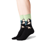 A pair of crew socks worn on a person's feet, featuring a black lower half and a light green upper half with a printed parody of the Mona Lisa wearing sunglasses. thumbnail