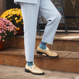 A person wearing light blue trousers, beige loafers with gold chain detailing, and blue patterned socks, standing on stone steps outdoors next to potted flowers.