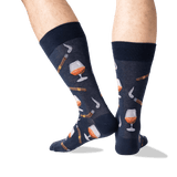 A pair of navy blue crew socks with a pattern of whiskey glasses, cigars, and smoking pipes, shown on a person's legs against a white background. thumbnail