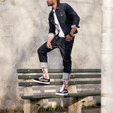 A man wearing a dark denim jacket, dark jeans, a white t-shirt, and black Vans sneakers with red and white socks, posing on a green wooden bench against a textured concrete wall. thumbnail