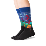 A pair of crew socks worn on a person's feet, featuring a colorful graphic of the Golden Gate Bridge and a trolley train in San Francisco on a blue background, with a black toe and heel section.