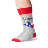 A pair of crew socks worn on a person's legs, featuring a heather gray body with a red cuff, red toe, and red heel. The socks display 'TEXAS' in navy blue text above a graphic of the state of Texas filled with the Texas flag and a cowboy wearing a sombrero.