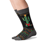 A pair of crew socks worn on a person's feet, featuring a black background with a green cactus in a red pot, wrapped in festive red and green geometric patterns, and topped with a yellow star.