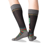 A pair of crew socks worn on a person's legs, featuring a dark charcoal gray base with a colorful geometric pattern in red, green, and teal, and a cactus motif on the side.
