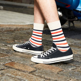 A person wearing navy low-top canvas sneakers with white stripes and cream, navy, and orange striped socks, standing on a cobblestone street. thumbnail