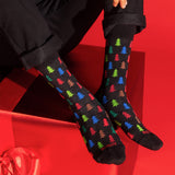 A person wearing black crew socks with a colorful Christmas tree pattern, posed against a bright red background with a red gift box.