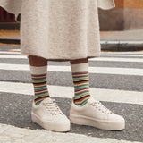 A person standing on a crosswalk wearing cream platform sneakers, a beige knit skirt, and white crew socks with multicolored horizontal stripes. thumbnail