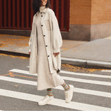 A woman walking across a city crosswalk wearing an oversized light beige button-front coat over a matching long-sleeve knit dress, with striped socks and white sneakers. thumbnail
