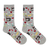 A pair of heather gray crew socks with an all-over sewing-themed pattern featuring black sewing machines, red hearts, yellow spools, scissors, and other colorful thread icons, shown against a plain white background. thumbnail