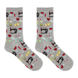 A pair of heather gray crew socks with an all-over sewing-themed pattern featuring black sewing machines, red hearts, yellow spools, scissors, and other colorful thread icons, shown against a plain white background.