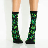 A pair of crew socks with a green cannabis leaf pattern on a black background, worn on a person's legs against a white studio backdrop.