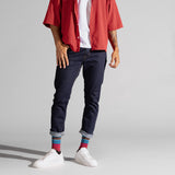 A model wearing dark blue slim-fit jeans with rolled cuffs, styled with a red striped short-sleeve button-up shirt, a white t-shirt, colorful striped socks, and white sneakers, set against a plain light gray studio background. thumbnail