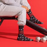 A person seated on a black exercise bench wearing black crew socks with white barbell graphics and red text reading 'PRACTICE SAFE SETS'. The scene is on a bright red studio background with dumbbells nearby.