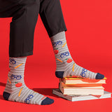 Gray striped socks with a school-themed pattern of blue glasses, yellow pencils, and red apples, worn with black pants and styled on a stack of books against a bright red background.
