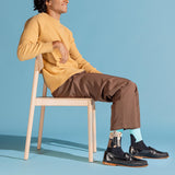A man sits on a light wood chair wearing a mustard yellow ribbed sweater, brown trousers, black loafers, and patterned socks against a solid light blue studio background.
