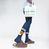 Person wearing blue jeans and brown shoes with a white tote bag featuring 'New York' text.