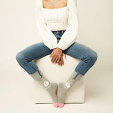 A model sits on a white textured cube sheep gray crew socks with a cute bunny graphic and pink toes, paired with blue jeans and a white long-sleeve top against a light beige background. thumbnail