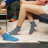 Gray ankle socks with a cat face design worn by a person sitting on a couch in a living room setting, alongside a blue knit slipper and another person's legs.