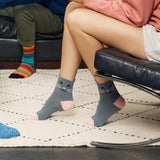 A person sitting on a black chair wearing gray socks with a cat face design and pink toes, with another person's legs visible in the background wearing colorful striped socks in a home setting.