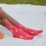 A person sitting outdoors on a white blanket, wearing bright pink crew socks with printed champagne bottle, champagne glasses, and orange slices. A glass of orange drink is visible at the lower left. thumbnail