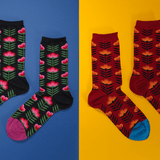 Two pairs of patterned crew socks displayed on a split blue and yellow background. The left pair is black with pink flowers and green leaves, and the right pair is dark red with orange and yellow leaves and blue toes. thumbnail