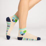 A pair of ankle socks worn on a model's feet, shown against a clean light gray studio background. The socks are heather beige with a colorful geometric heart design in blue, green, and navy on the ankle, striped bands in blue, green, and white on the sides, and solid navy heels, toes, and cuffs.