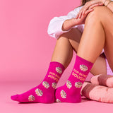 A person seated against a pink studio background wearing bright pink crew socks printed with cupcake illustrations and the phrase “DEM BUNS DOUGH.”.