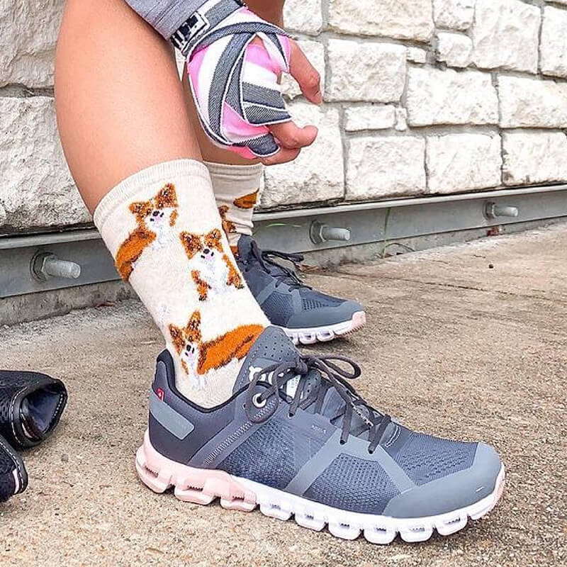Person wearing gray athletic shoes with white soles, colorful socks with a corgi dog design, and pink and gray gloves on a stone pavement.