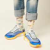 A person wearing cuffed blue jeans, patterned cream socks with text and fish illustrations, and chunky blue, white, and yellow sneakers against a plain light beige studio background.