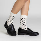 A person's legs wearing cream-colored crew socks with a black heart pattern, paired with black leather penny loafers, shown against a light gray studio background. thumbnail