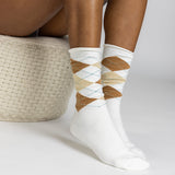 White crew socks with an argyle pattern in brown, beige, and light blue, worn by a model sitting on a woven straw stool.