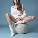 Woman sitting on a woven stool wearing white  sheer slouch socks against a blue background thumbnail