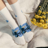 White socks with blue floral patterns held by a person next to a bouquet of yellow flowers on a textured surface. thumbnail
